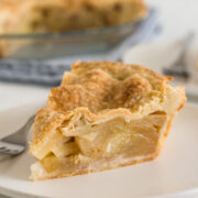 close-up side view of a slice of apple pie on a white plate with a fork and with the rest of the pie in the background
