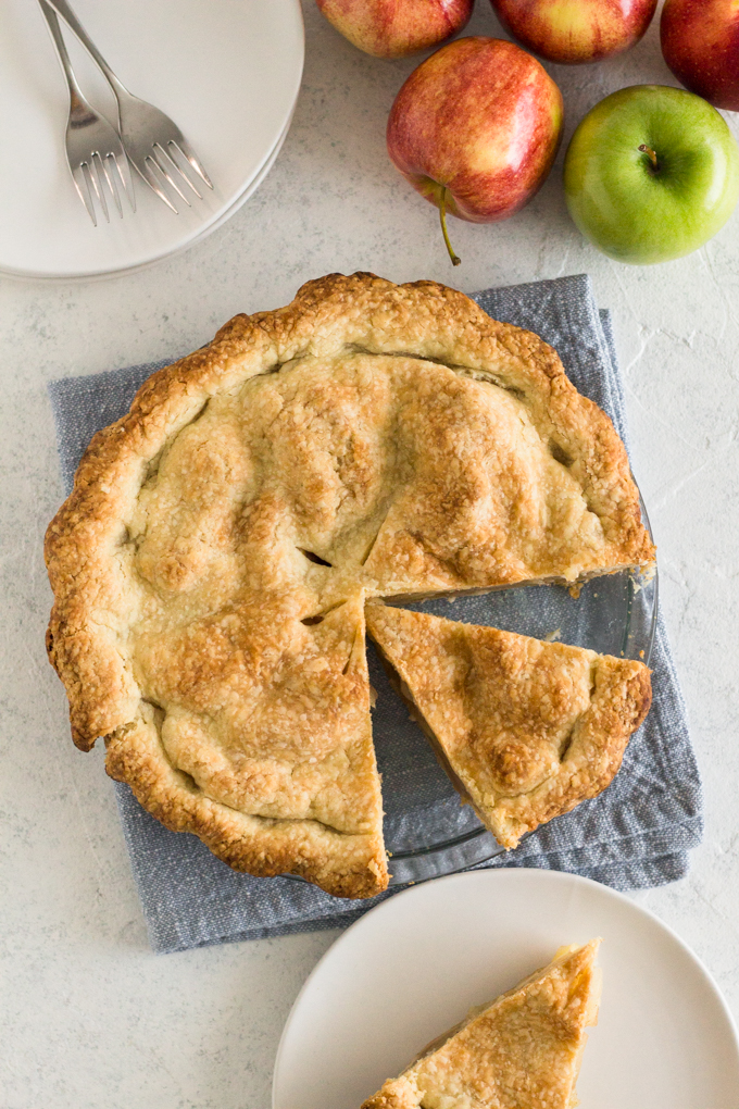 overhead view of a sliced apple pie on a blue cloth napkin with apples and white plates