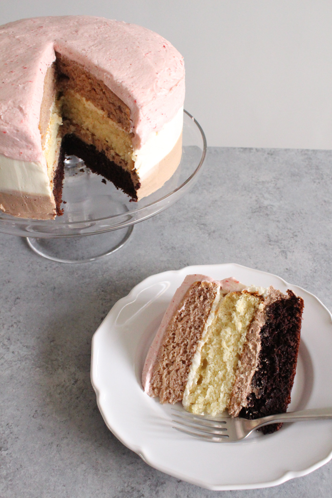 angled view of a slice of neapolitan cake on a white plate next to cake on a glass cake stand