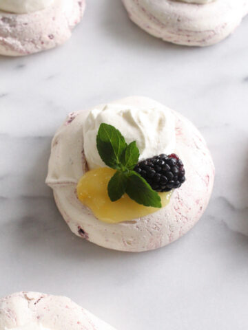 overhead view of meringue topped with lemon curd, whipped cream, blackberry, and mint sprig on a marble surface