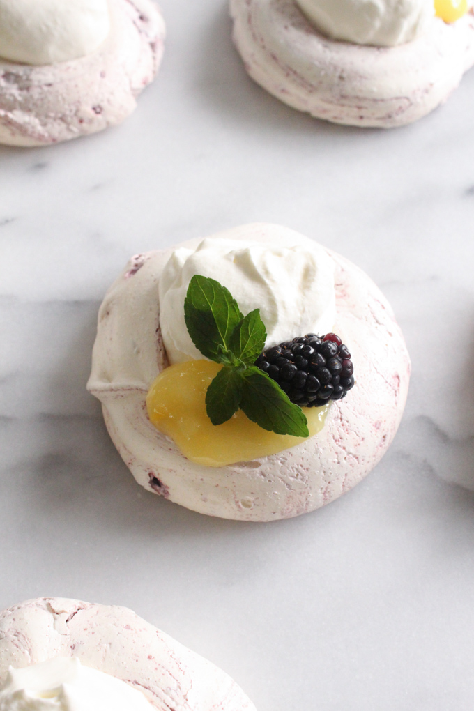 overhead view of meringue topped with lemon curd, whipped cream, blackberry, and mint sprig on a marble surface