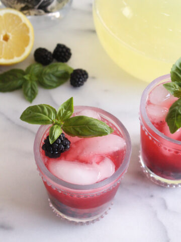 angled view of blackberry lemonade in glasses garnished with fresh basil sprigs on a marble surface
