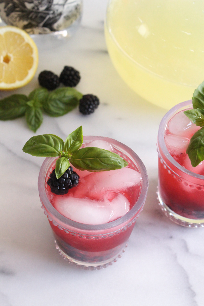 angled view of blackberry lemonade in glasses garnished with fresh basil sprigs on a marble surface