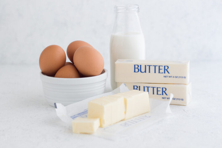 butter, brown eggs, and a glass of milk on a white surface