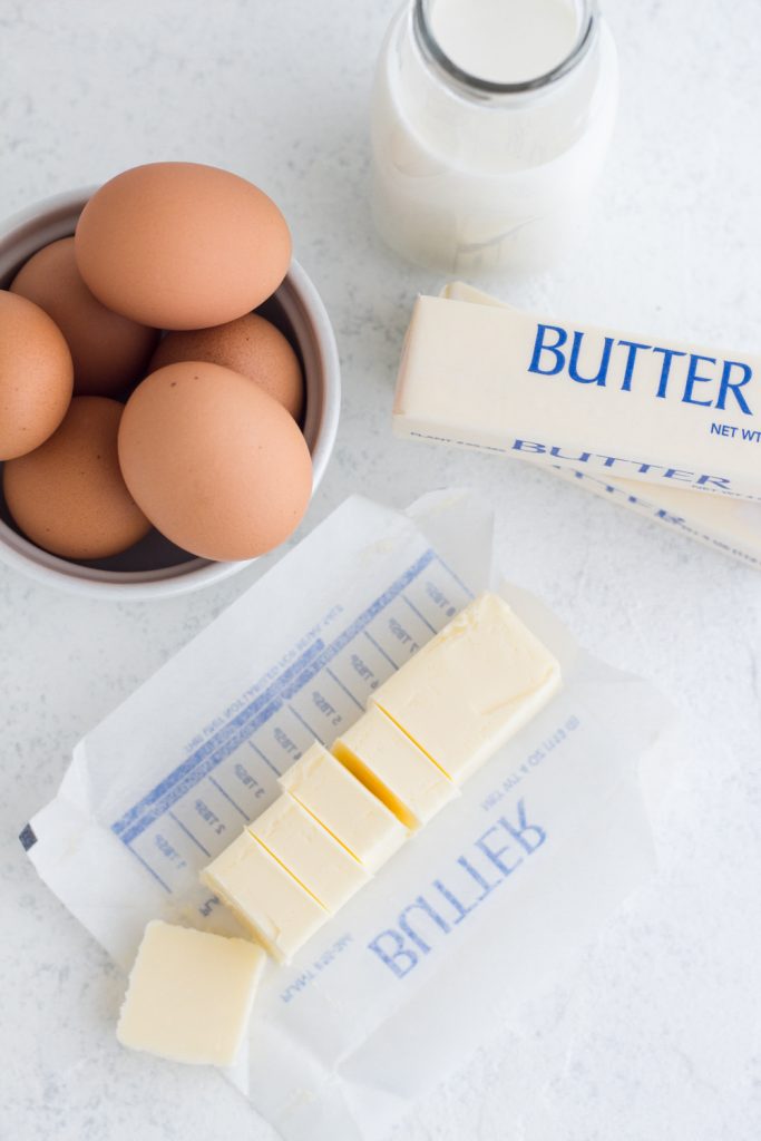 overhead view of sliced butter, brown eggs, and a glass of milk on a white surface