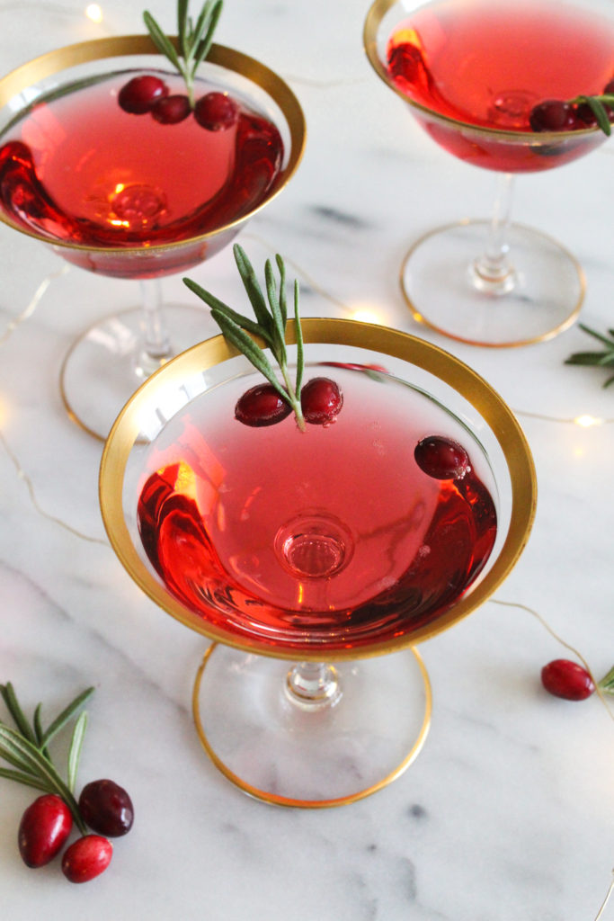 angled view of cranberry champagne cocktail in gold-rimmed coupe glasses on a marble surface