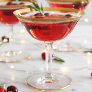 side view of 3 cranberry champagne cocktail in gold-rimmed coupe glasses on a marble surface