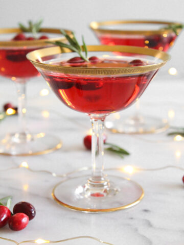 side view of 3 cranberry champagne cocktail in gold-rimmed coupe glasses on a marble surface