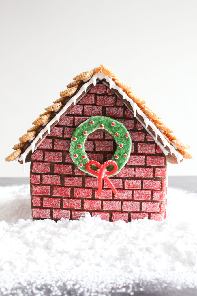 Gingerbread house with red "brick" walls and a wreath decorated with green sugar and red candies.