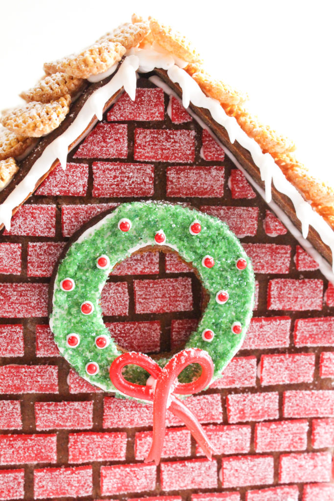 Close up view of gingbread house with red candy "brick" walls and a wreath decorated with green sugar and red candies.