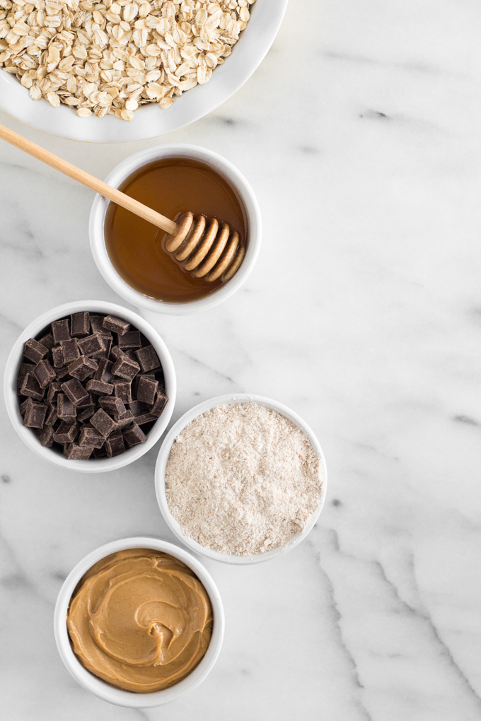 overhead view of breakfast cookie ingredients - oats, honey, dark chocolate chunks, whole wheat flour, and peanut butter - on a marble surface