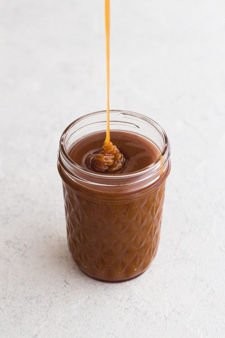 angled view of homemade salted caramel sauce being drizzled into a glass jar on a white surface
