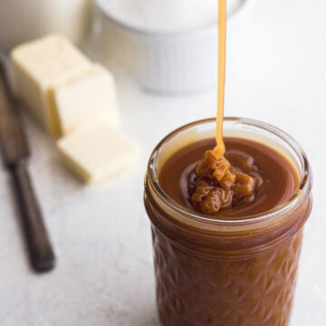 angled view of homemade salted caramel sauce being drizzled into a glass jar on a white surface with sugar, butter, and cream in the background