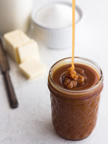 angled view of homemade salted caramel sauce being drizzled into a glass jar on a white surface with sugar, butter, and cream in the background
