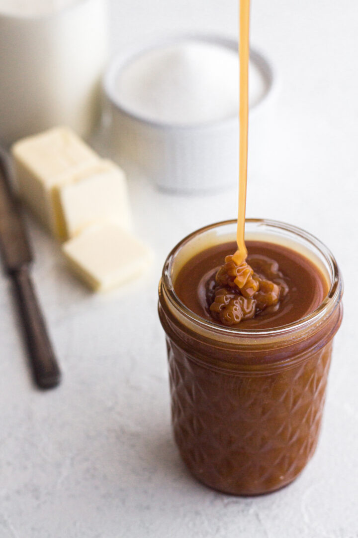angled view of homemade salted caramel sauce being drizzled into a glass jar on a white surface with sugar, butter, and cream in the background