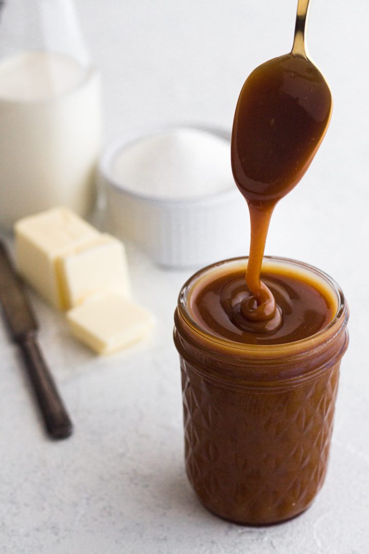 angled view of homemade salted caramel sauce being drizzled from a gold spoon into a glass jar on a white surface with sugar, butter, and cream in the background