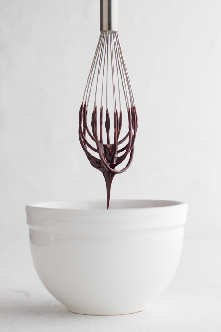side view of chocolate ganache dripping from a metal whisk into a white bowl on a white background