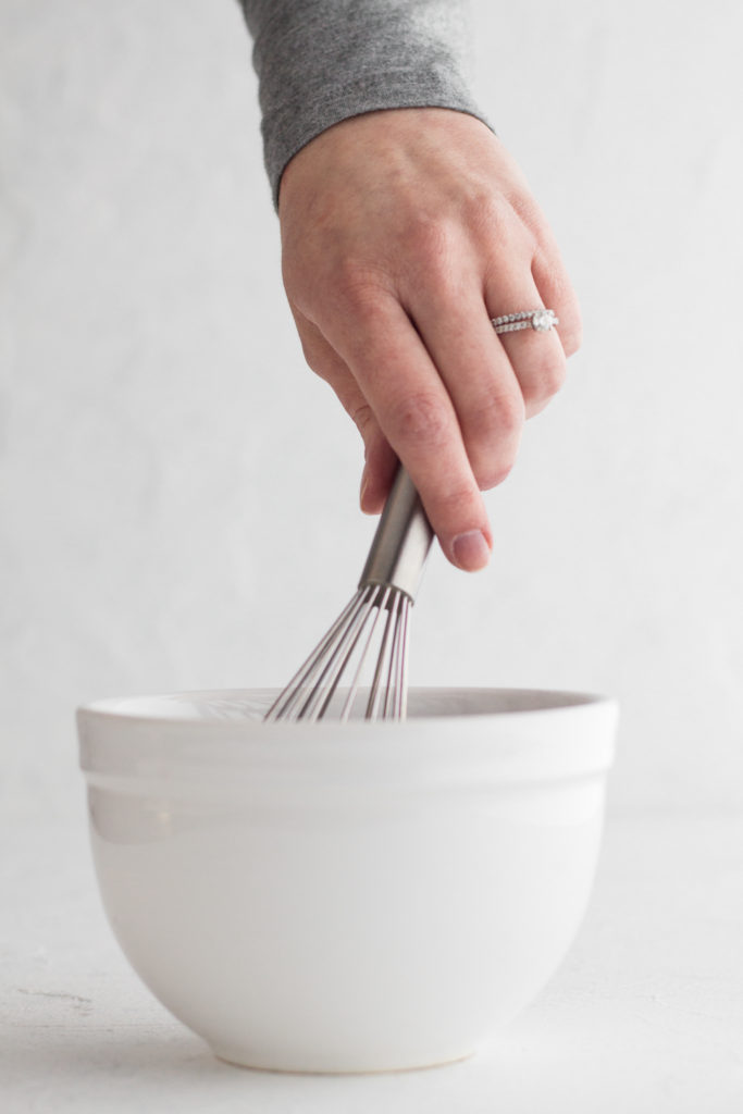 side view of hand holding a metal whisk above a white bowl on a white background