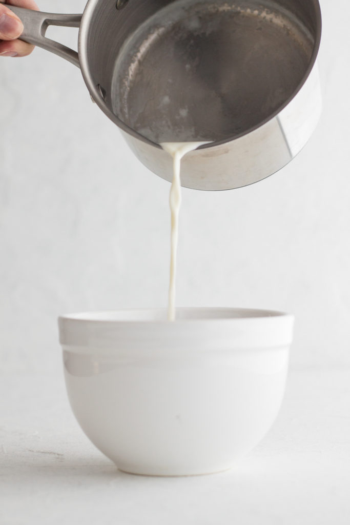 side view of cream being poured from a metal saucepan into a white bowl on a white backgroud
