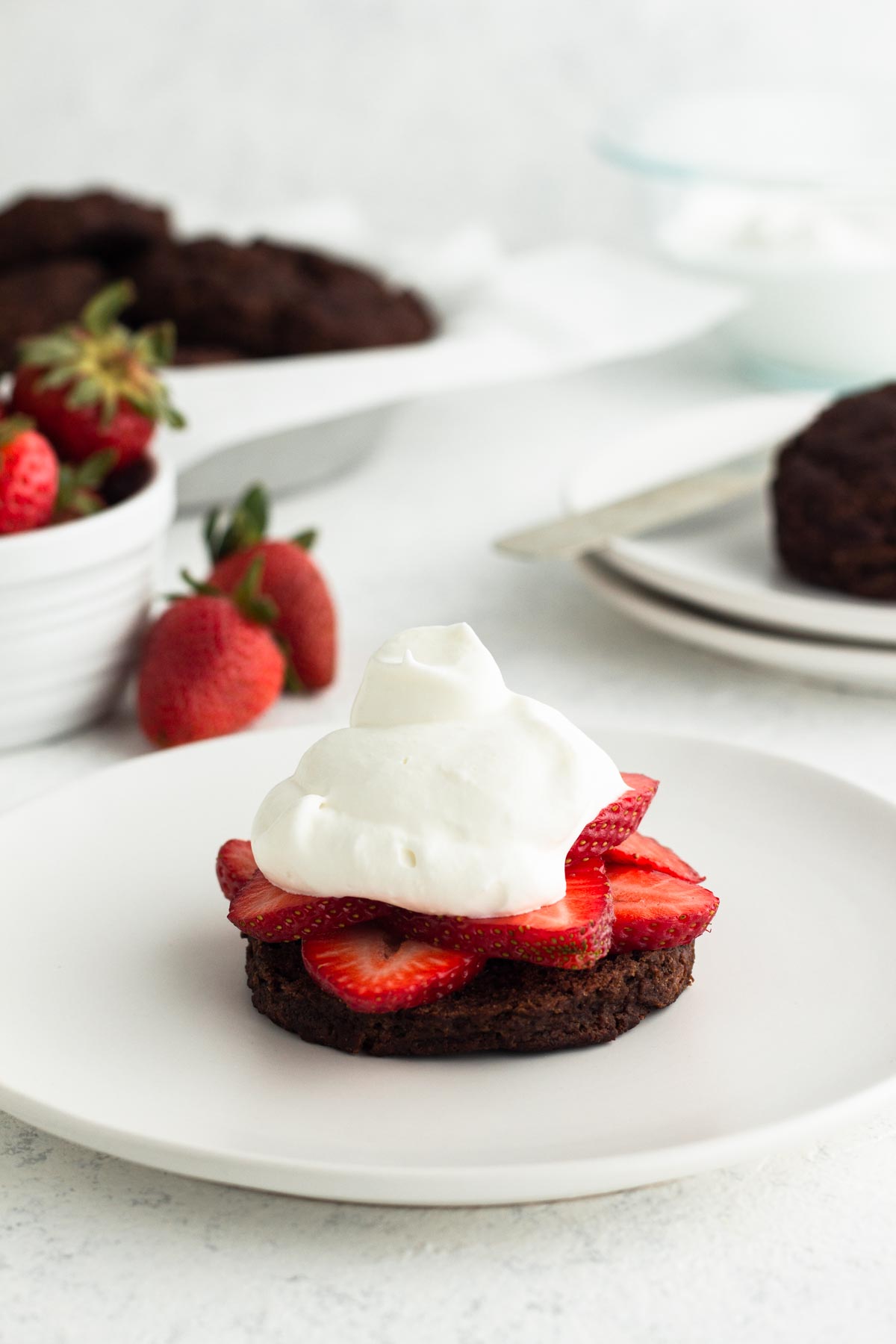 side view of a chocolate buttermilk biscuit topped with fresh strawberries and homemade whipped cream on a white plate