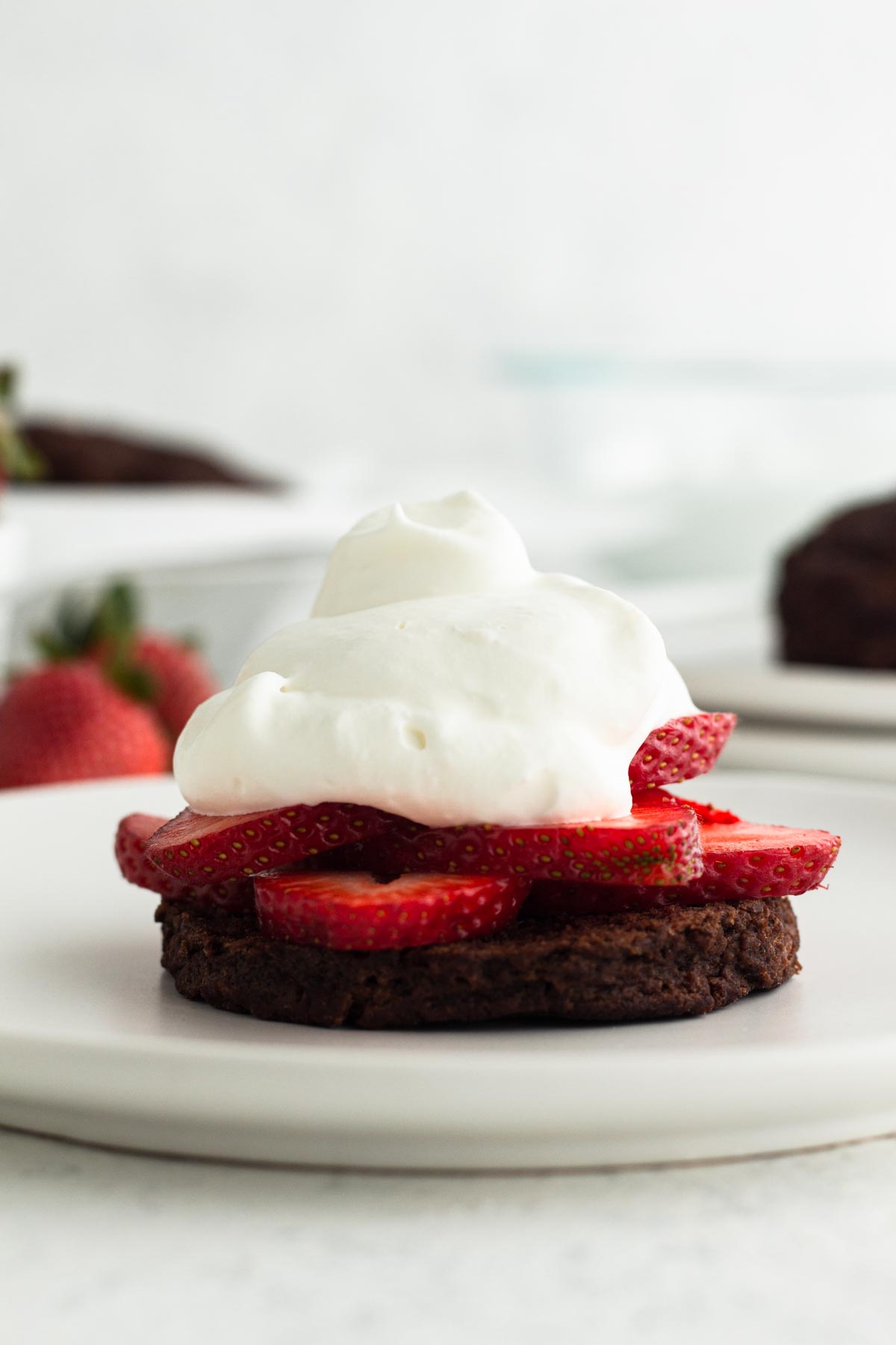 close up side view of a chocolate buttermilk biscuit topped with fresh strawberries and homemade whipped cream on a white plate