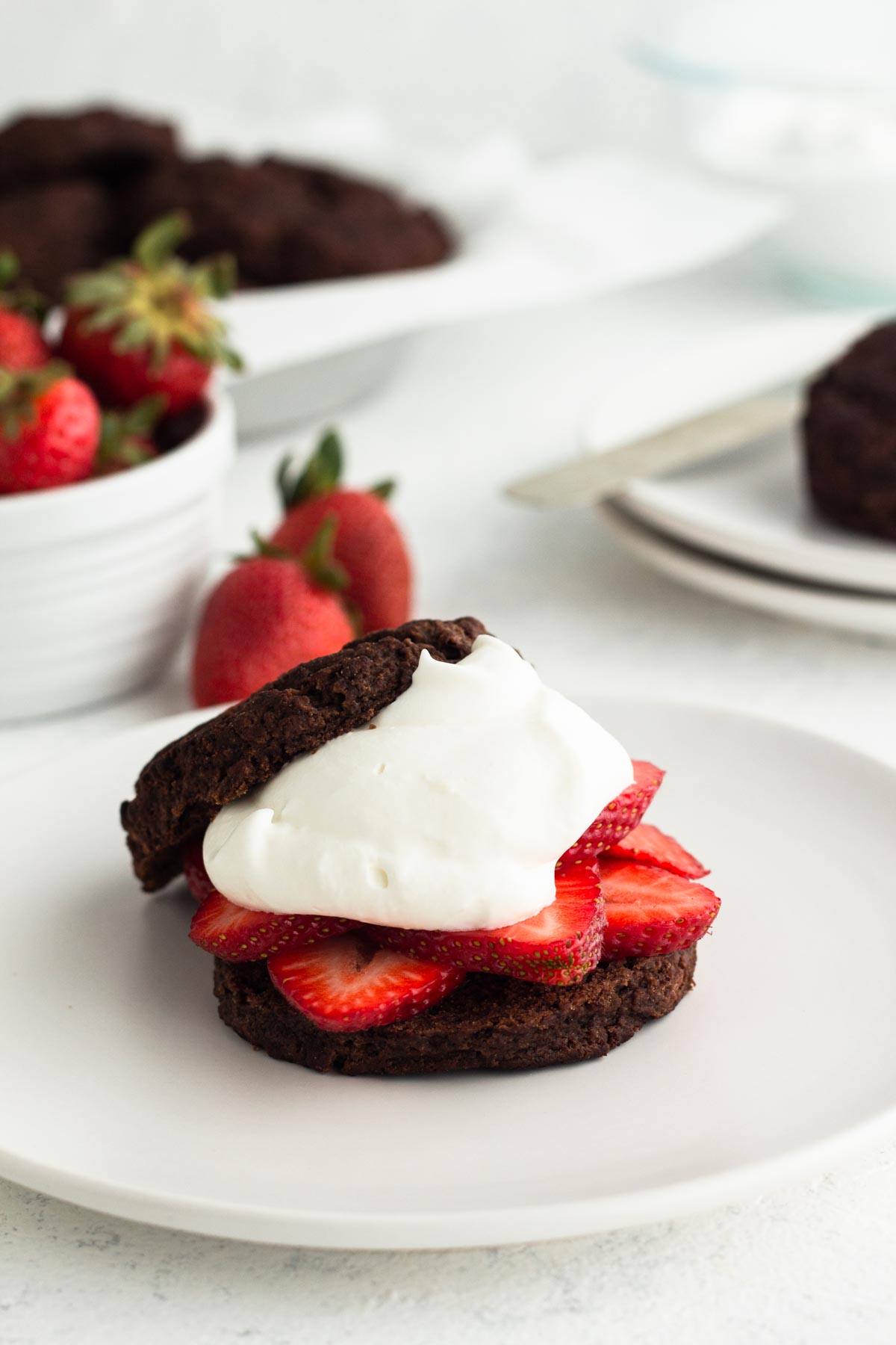 angled side view of a chocolate buttermilk biscuit topped with fresh strawberries and homemade whipped cream on a white plate