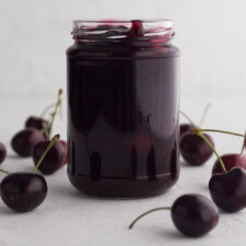 side view of homemade cherry sauce in a glass jar surrounded by fresh cherries on a white surface