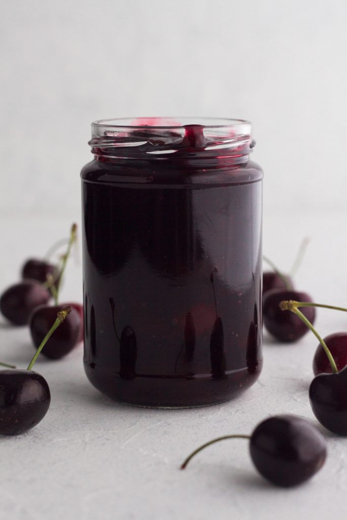 side view of homemade cherry sauce in a glass jar surrounded by fresh cherries on a white surface