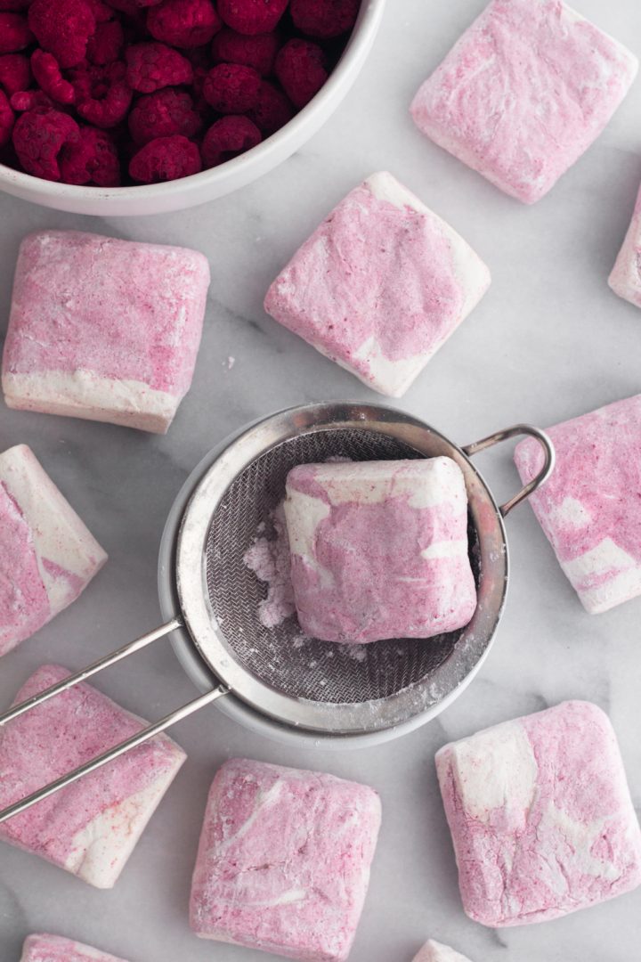 overhead view of homemade raspberry swirl marshmallows being dusted with confectioners sugar on a marble surface