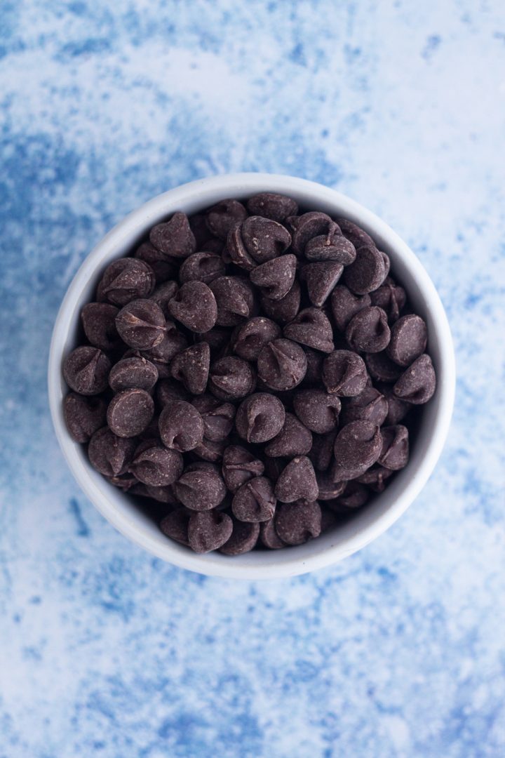 overhead view of chocolate chips in a white ramekin on a blue surface