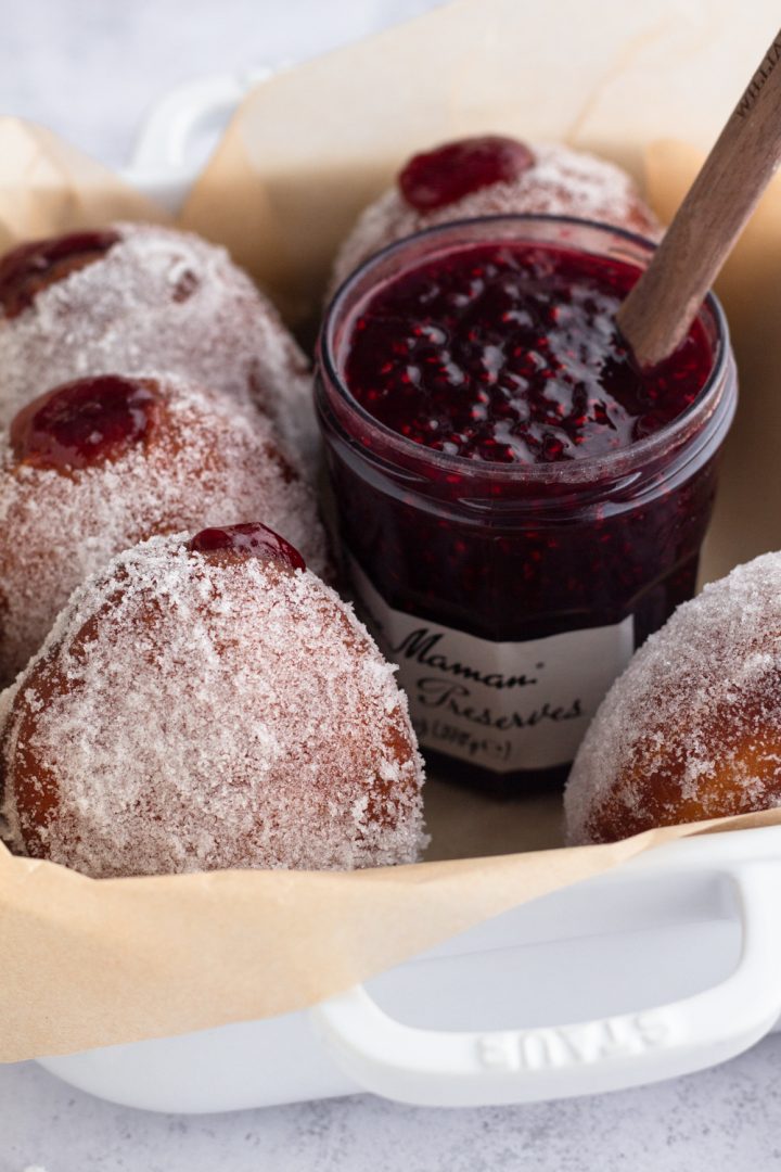 angled view of brioche jelly doughnuts and a jar of raspberry jam in a white dish with brown parchment paper