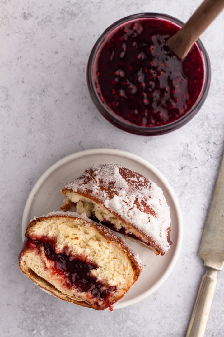 overhead view of a brioche jelly doughnut cut in half on a white plate with a jar of raspberry jam