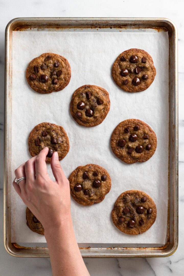 overhead view of chocolate chip cookies on a baking sheet with a hand adding chocolate chips on top of a cookie