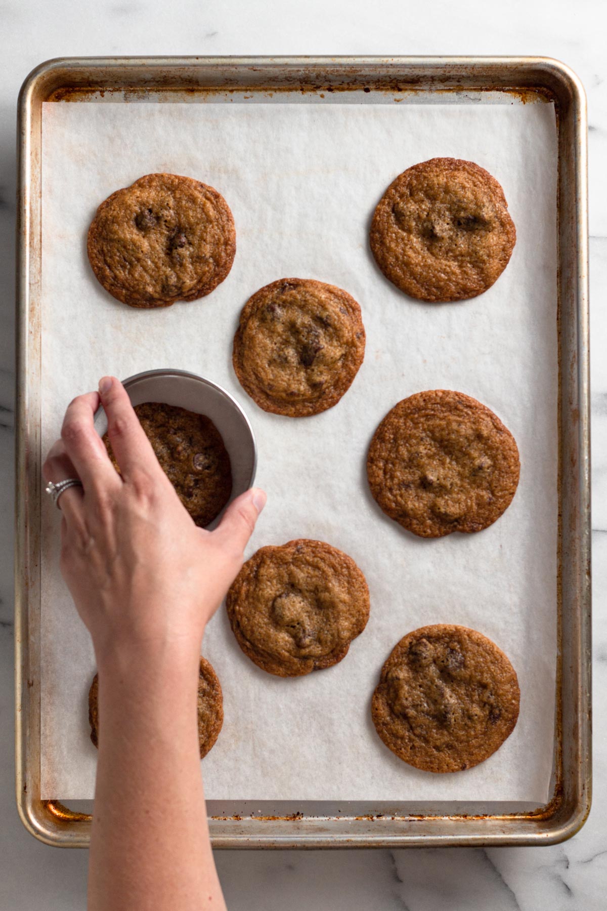 overhead view of baked chocolate chip cookies on a baking sheet with a hand holding a round cookie cutter