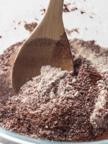 Flour and cocoa being stirred together in a glass bowl with a wooden spoon.
