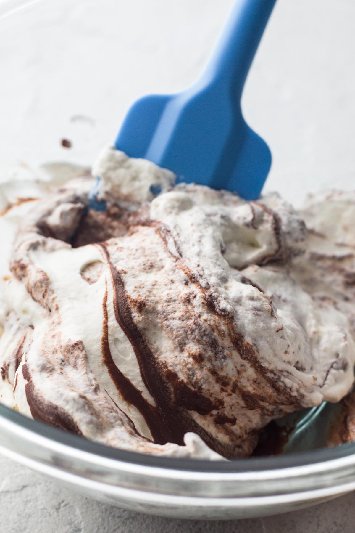 Whipped cream and melted chocolate being folded together in a glass bowl with a blue rubber spatula.