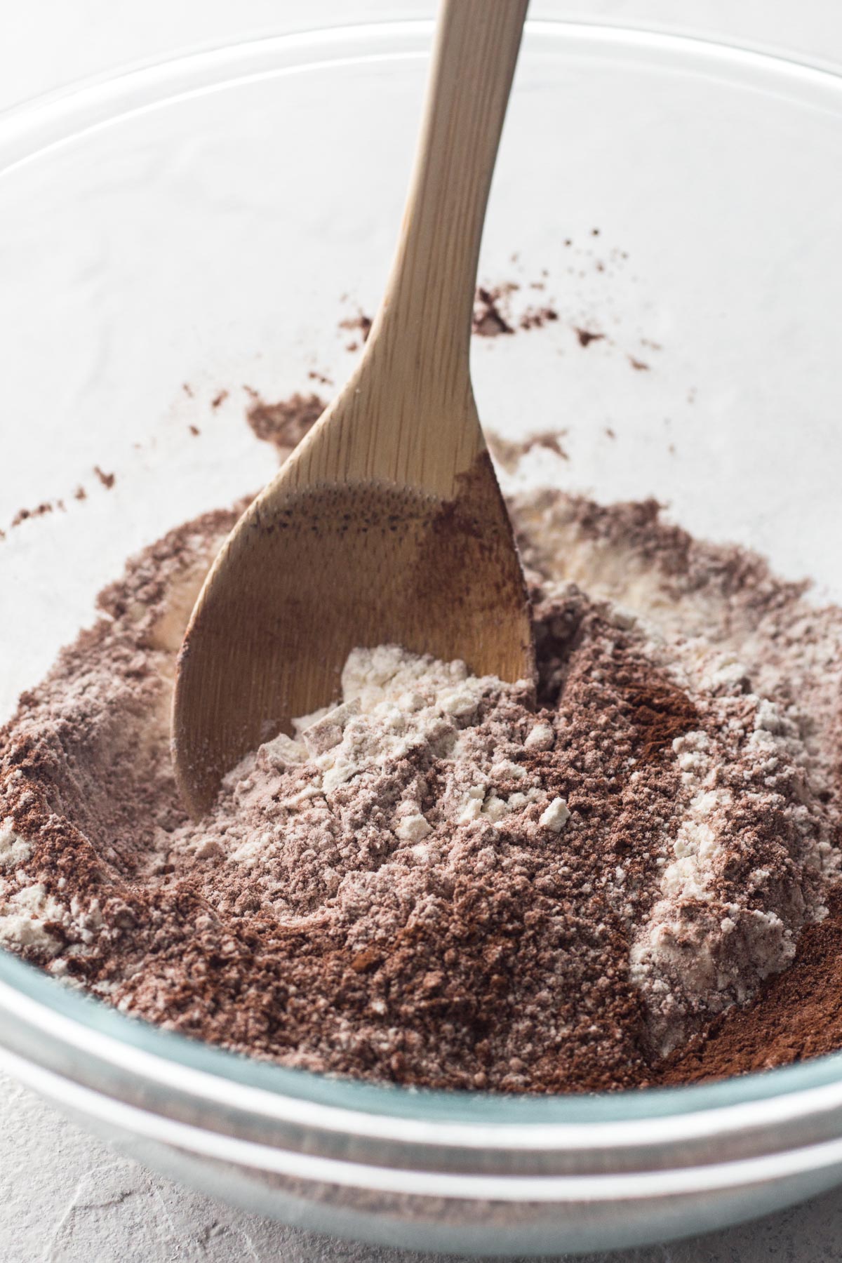 Flour and cocoa being stirred together in a glass bowl with a wooden spoon.