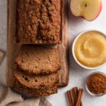 Sliced loaf on a wooden cutting board next to apples, applesauce, and cinnamon.