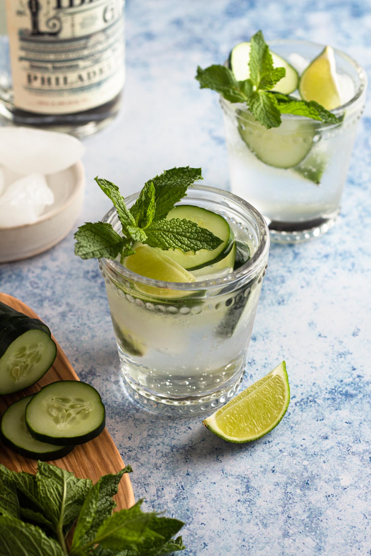 Gin and tonic garnished with a lime wedge, cucumber slices, and a sprig of mint on a speckled blue surface.