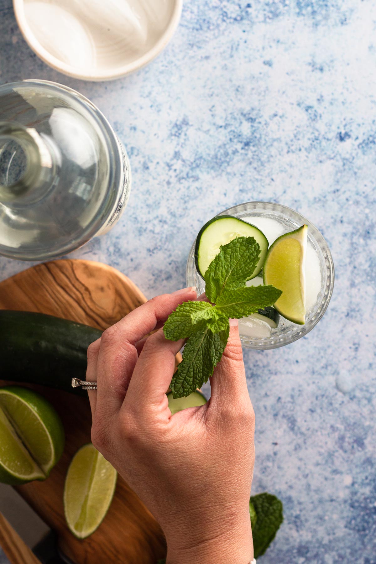 Hand adding a mint sprig to a rocks glass garnished with a lime wedge and cucumber slice.