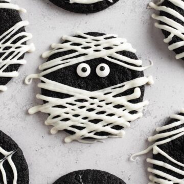 Overhead view of black cocoa cookies decorated like a mummy with white chocolate on a gray surface.