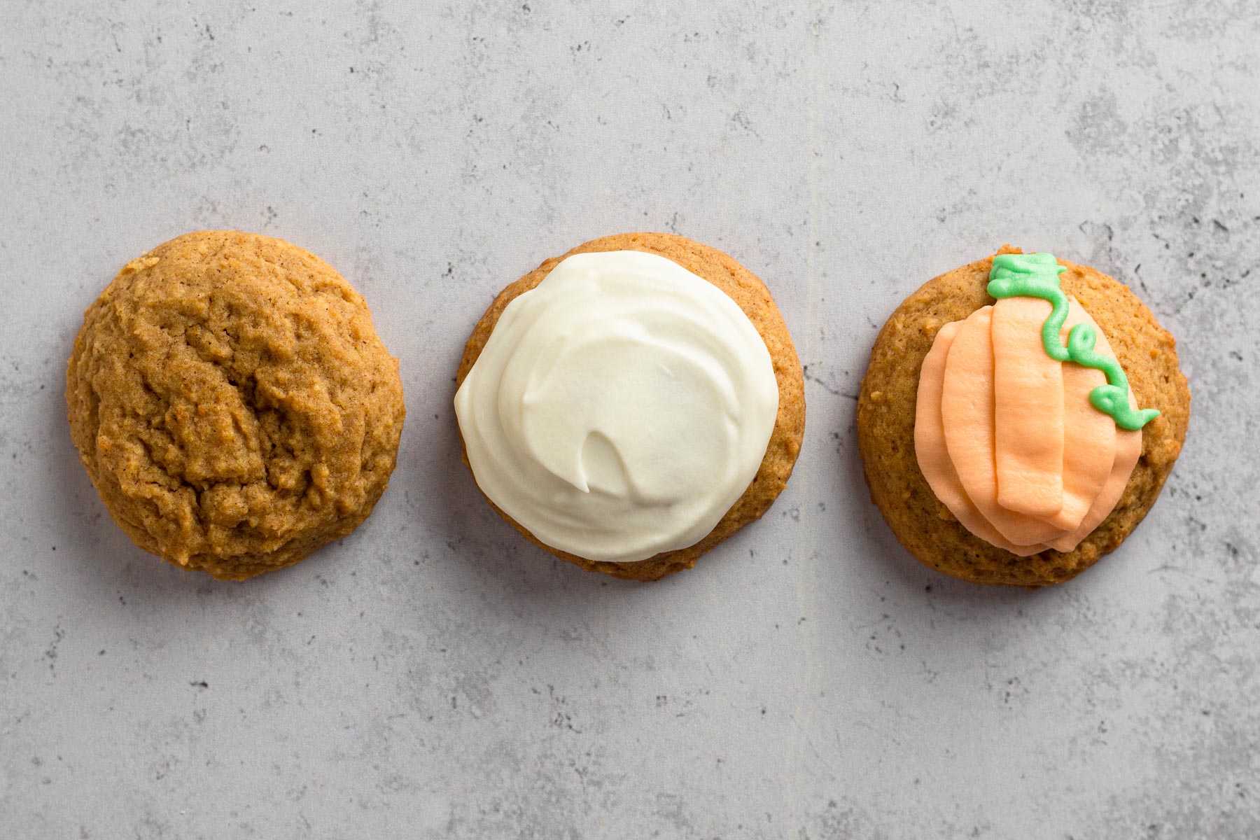 Three pumpkin cookies - one plain, one frosted, and one decorated - on a gray surface.
