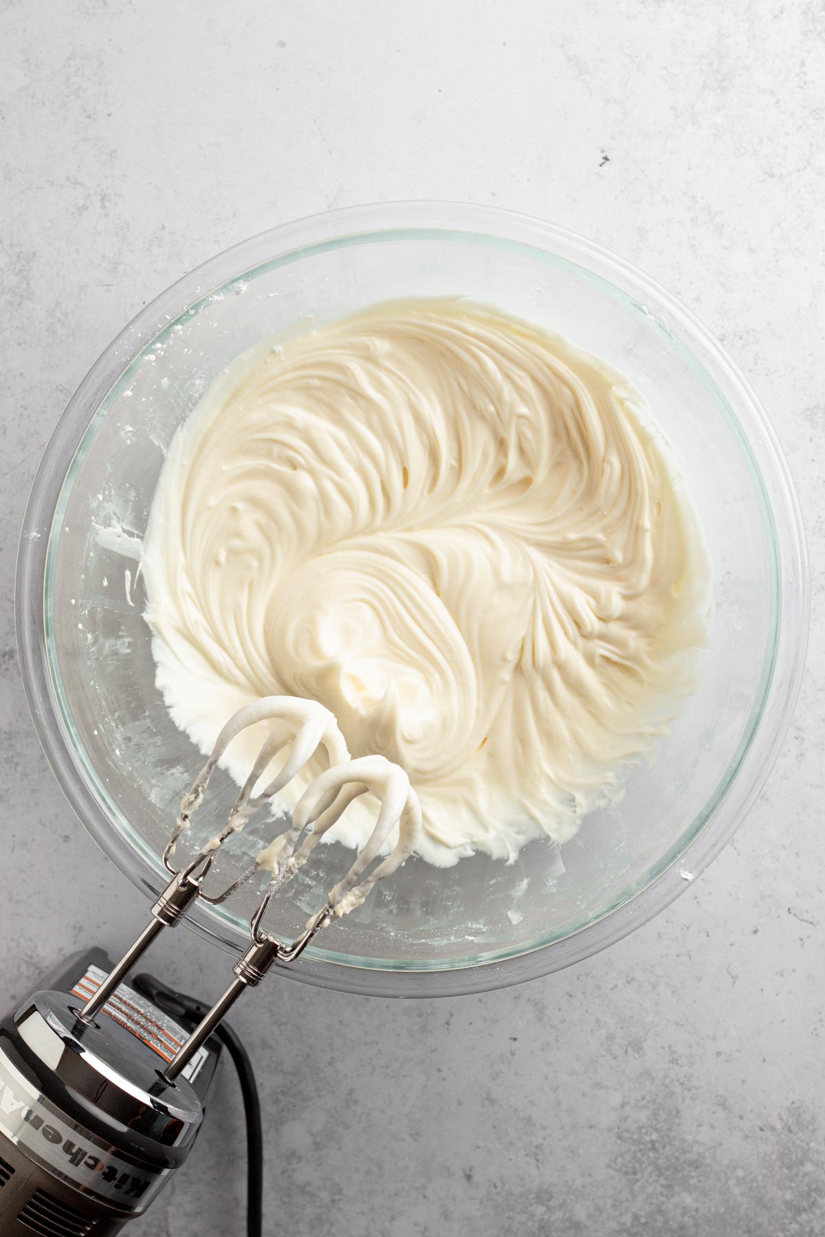 Cream cheese frosting in a glass bowl with an electric hand mixer.