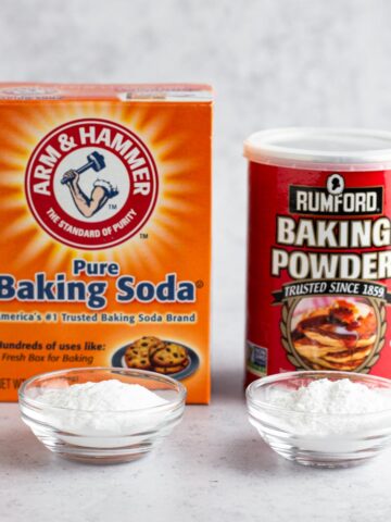 A box of baking soda and a can of baking powder next to small glass bowls of both leaveners.