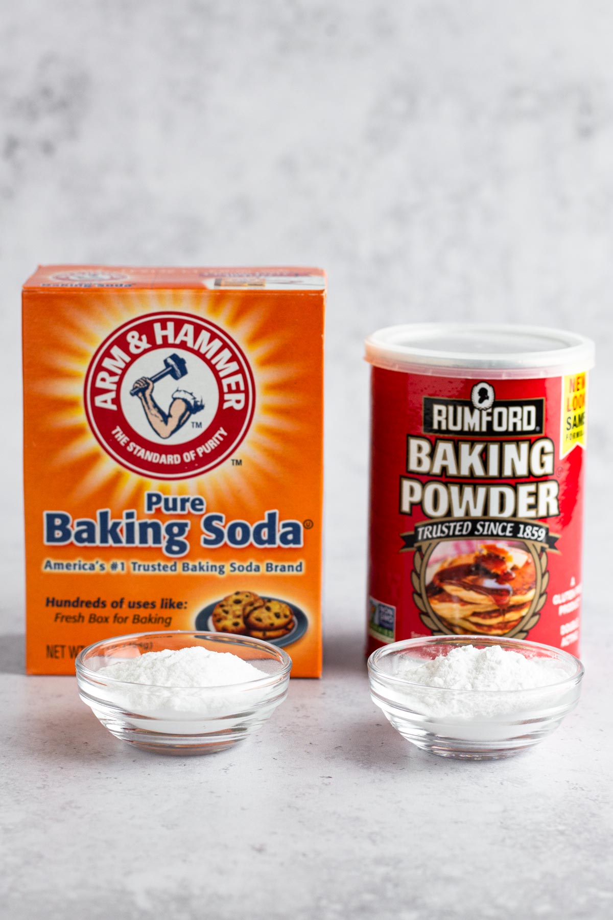 A box of baking soda and a can of baking powder next to small glass bowls of both leaveners.