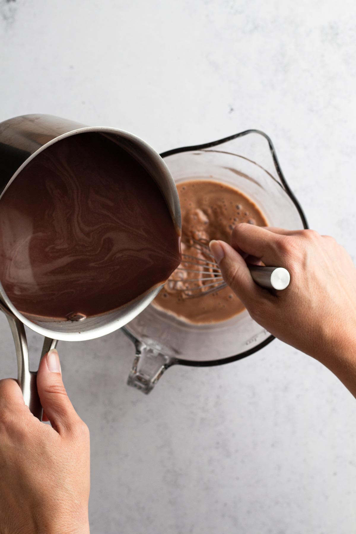 Hand pouring hot cocoa mixture from saucepan into glass bowl to temper egg mixture.