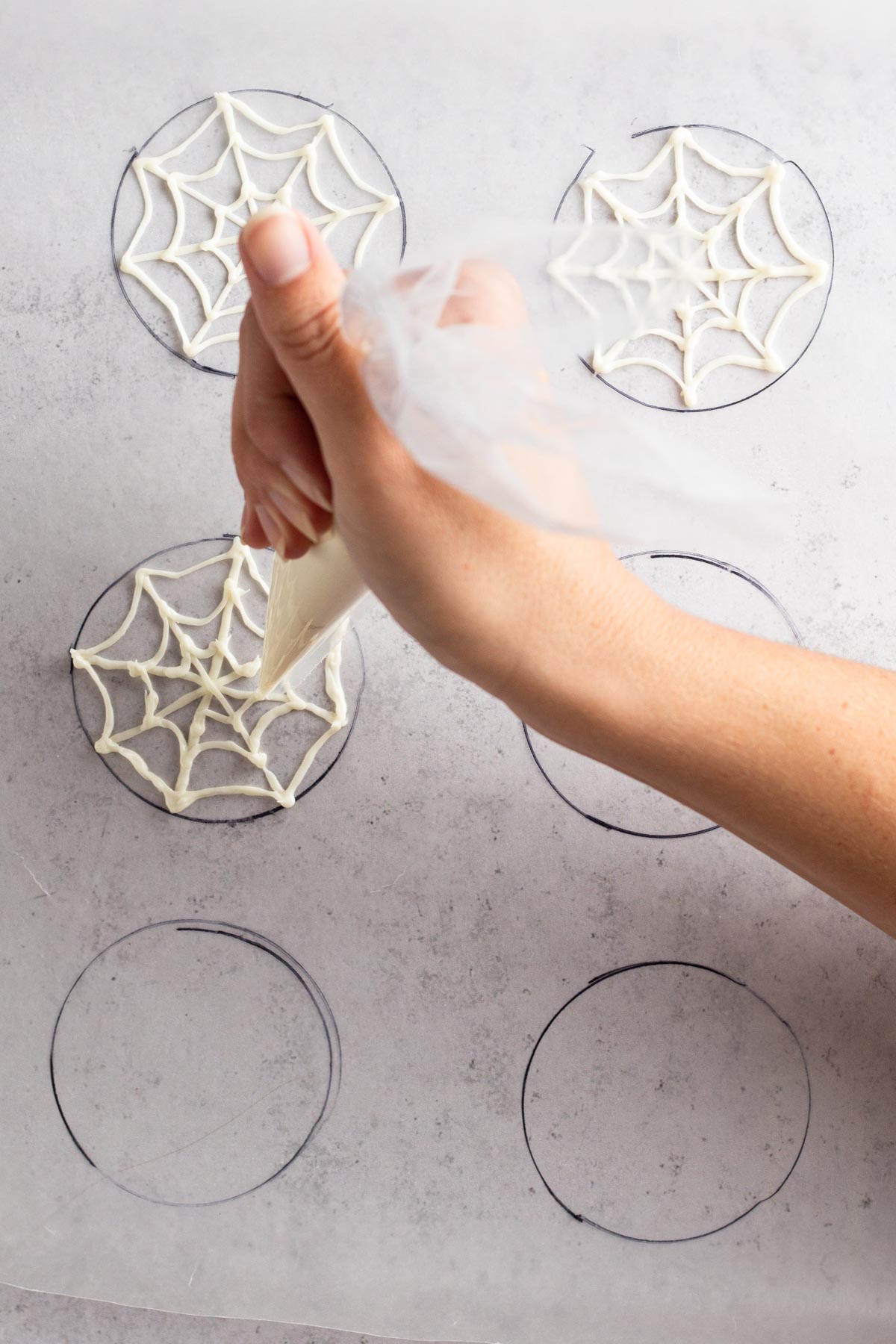 Hand piping swooped lines of melted white chocolate onto wax paper to complete a spiderweb pattern.