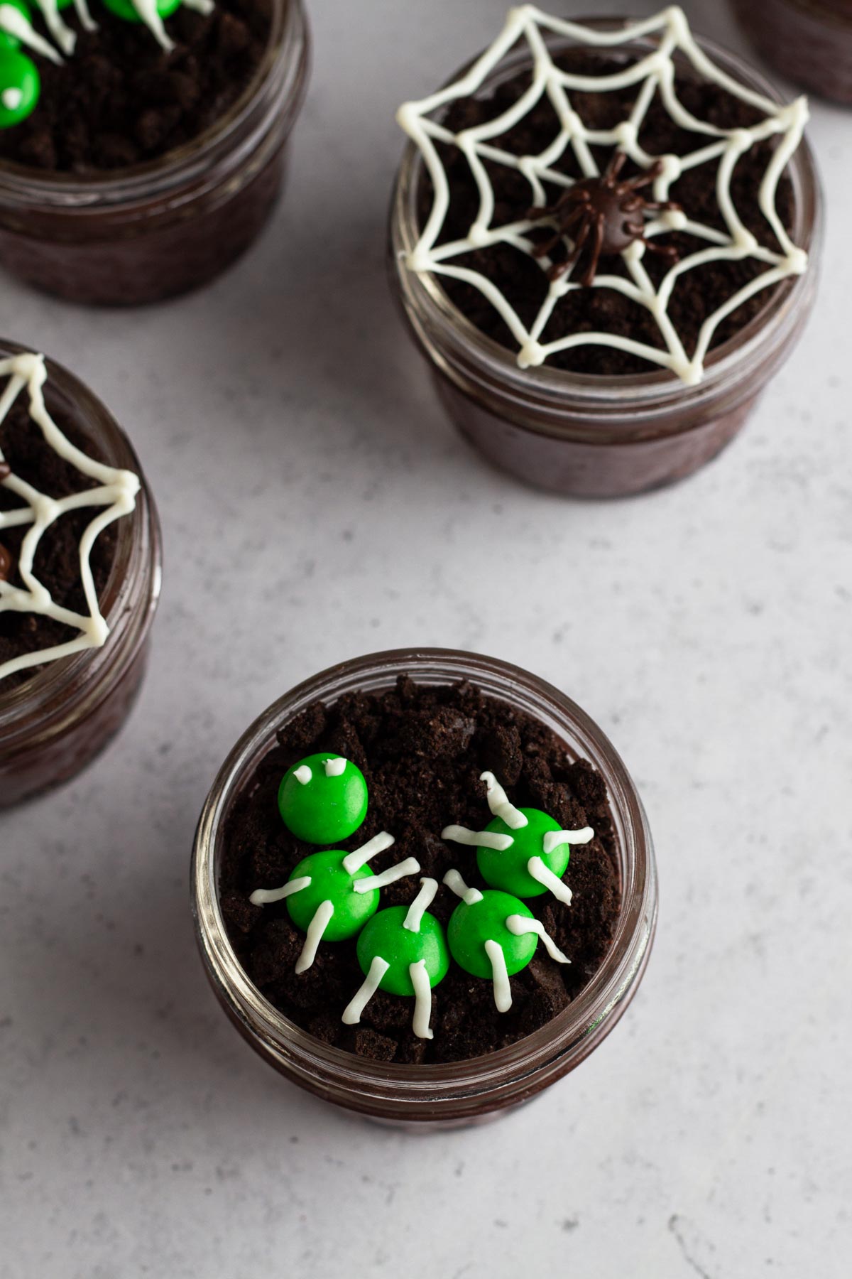 Dirt pudding cups in glass jars topped with chocolate and M&M bugs on a gray surface.
