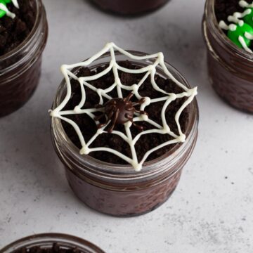 Dirt pudding cup in a glass jar topped with a white chocolate spiderweb and M&M spider.