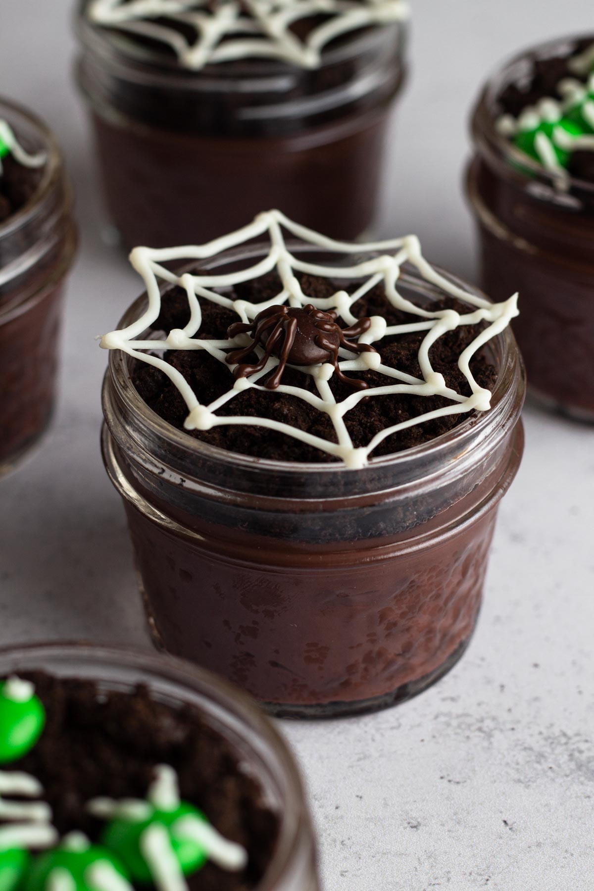 Dirt pudding cup in a glass jar topped with a white chocolate spiderweb and M&M spider.